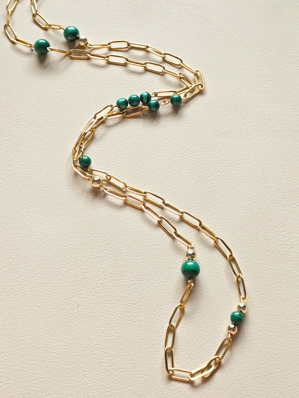 Coral Heat Malachite and Gold Paperclip Chain 36-inch necklace laid out in an elegant S-curve on a neutral textured background, showing grouped green gemstone beads and gold accents.